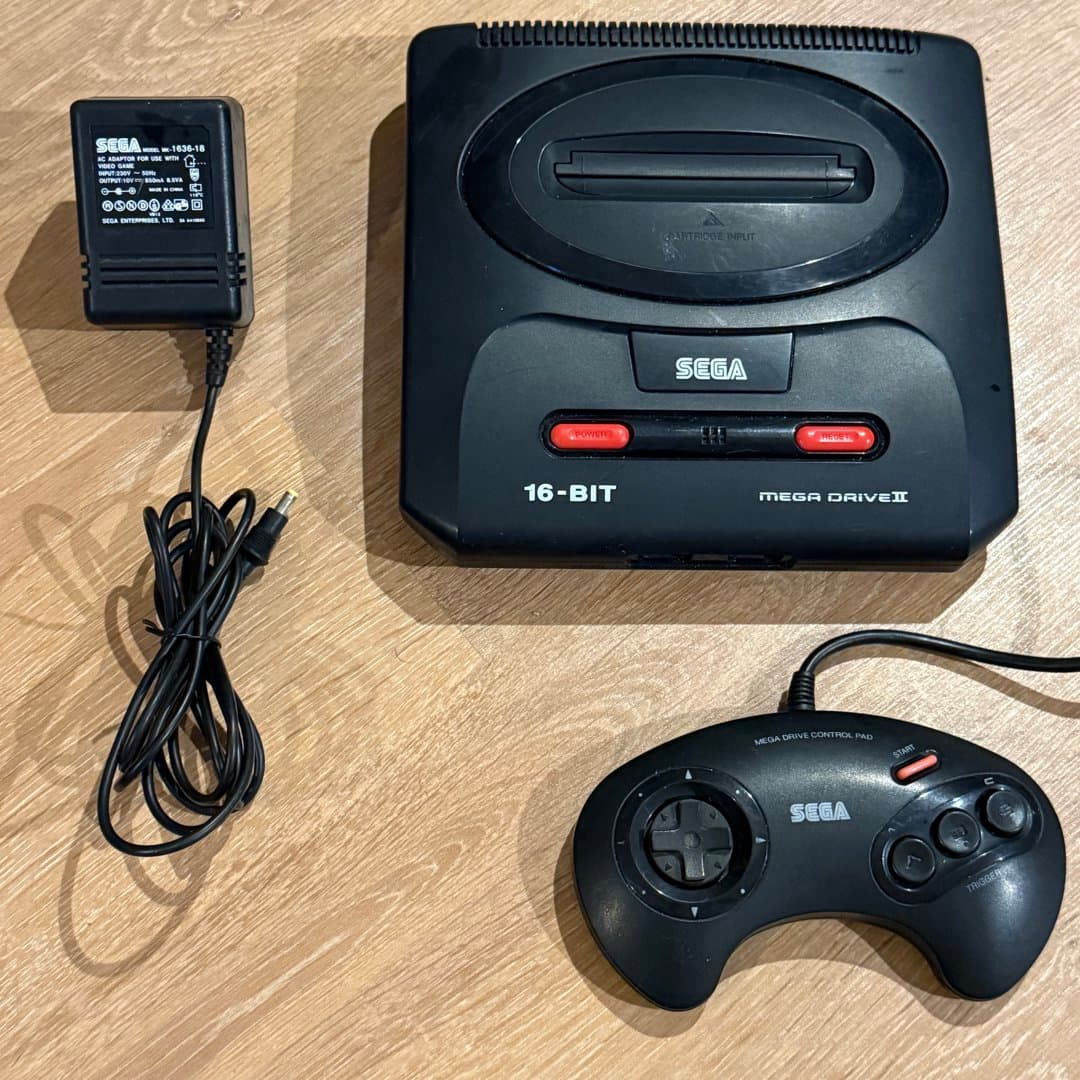 A vintage Sega Mega Drive II 16-bit console with its original controller and power adapter.